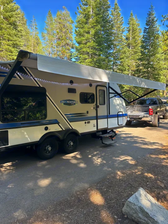 RV rental travel trailer with extended awning and lights, hitched to a pickup truck at a sunny campsite among tall pine trees.. Jayco Jay Flight 2018