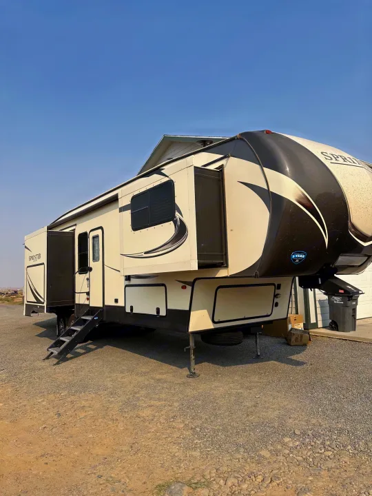This cream and black Sprinter fifth-wheel RV rental, with extended slide-outs and steps, is parked on a gravel lot.. Keystone Sprinter 2018
