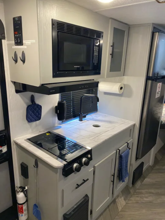 View of an RV rental kitchen showing a microwave above a stovetop, a sink, and light grey cabinets, all designed for compact, modern living.. Forest River Salem FSX 2022