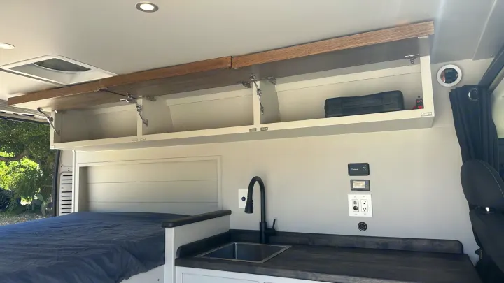 Explore this RV rental's cozy interior: bed, kitchen sink, open storage, and power panel. Perfect camper van.. RAM Promaster 2024