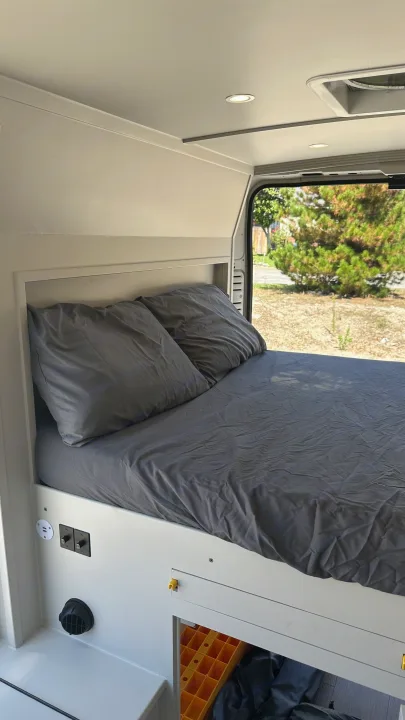 A comfortable bed with dark gray bedding and integrated charging ports inside a bright RV rental. Scenic outdoor view.. RAM Promaster 2024