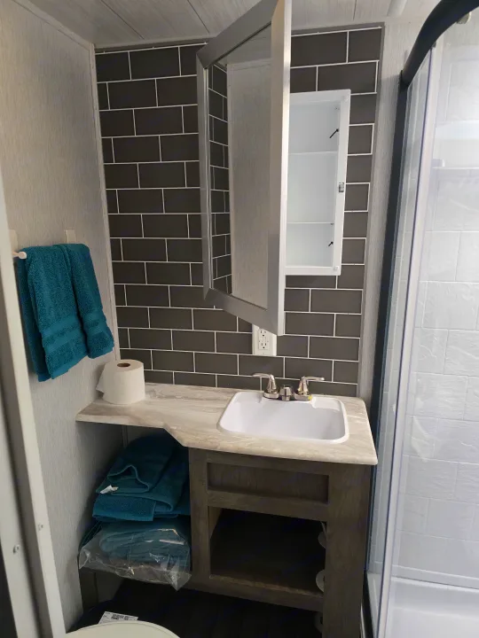 RV rental bathroom featuring a vanity sink, open medicine cabinet, dark tiled backsplash, teal towels, and a shower.. Forest River Wildwood 2022