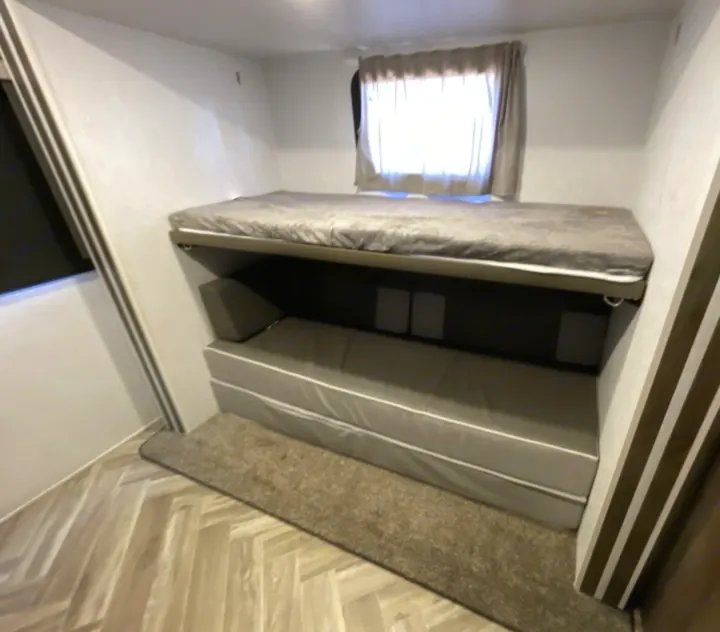 Inside an RV rental: a cozy bunkhouse with gray bunk beds, bright window, and light wood herringbone floor.. Forest River Wildwood 2022