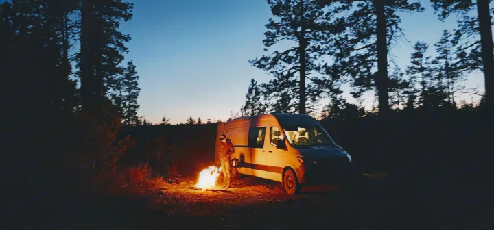 Twilight camping with an RV rental: A person by a roaring campfire next to a camper van in a serene forest at dusk.. Sprinter Mercedes 2018