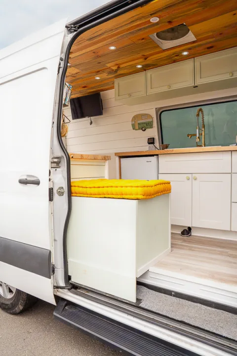 Inside a custom RV rental camper van: wooden ceiling, kitchenette, TV, and a yellow-cushioned bench visible via the open door.. Mercedes-Benz Sprinter 2019