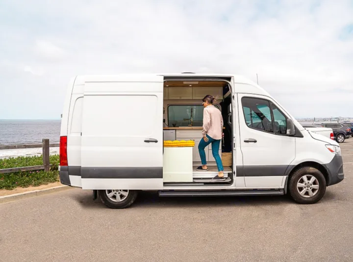 Woman stepping into a custom white camper van, an RV rental, parked by the ocean. Perfect for road trips.. Mercedes-Benz Sprinter 2019