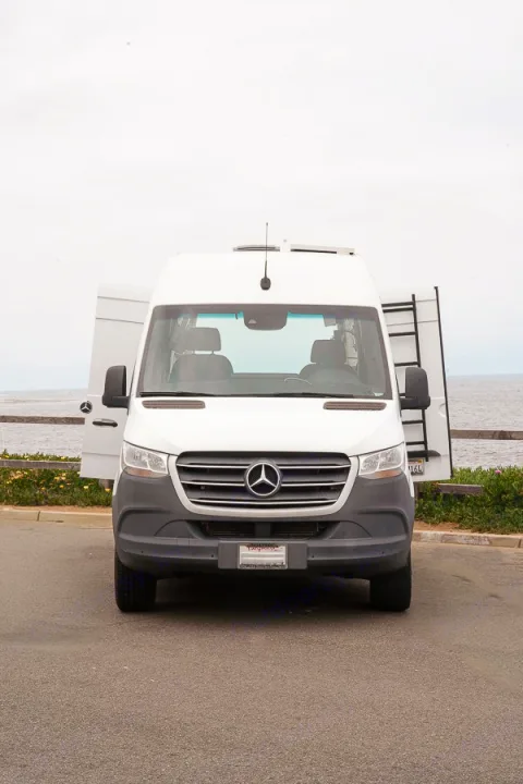 Front view of a white Mercedes-Benz Sprinter rv rental van. Its rear doors are open, revealing a ladder, parked by the ocean.. Mercedes-Benz Sprinter 2019
