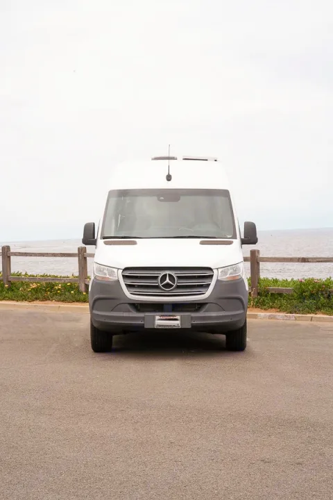 Front view of a white Mercedes-Benz Sprinter camper van, an RV rental, parked on asphalt by the ocean.. Mercedes-Benz Sprinter 2019