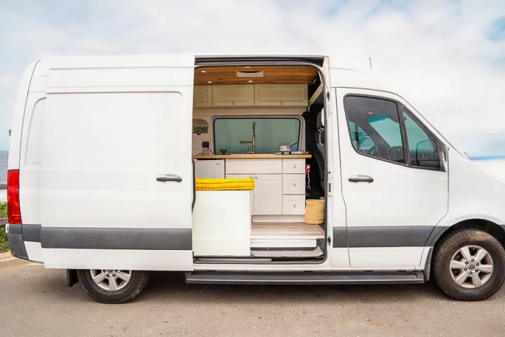 This custom white camper van, perfect for an RV rental, shows its open side door revealing a stylish kitchen and cozy interior setup for travel.. Mercedes-Benz Sprinter 2019