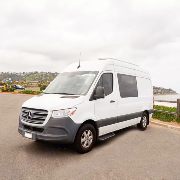 A white Mercedes-Benz Sprinter high-roof van, converted for camping and available for rv rental, parked by a scenic coastline.. Mercedes-Benz Sprinter 2019