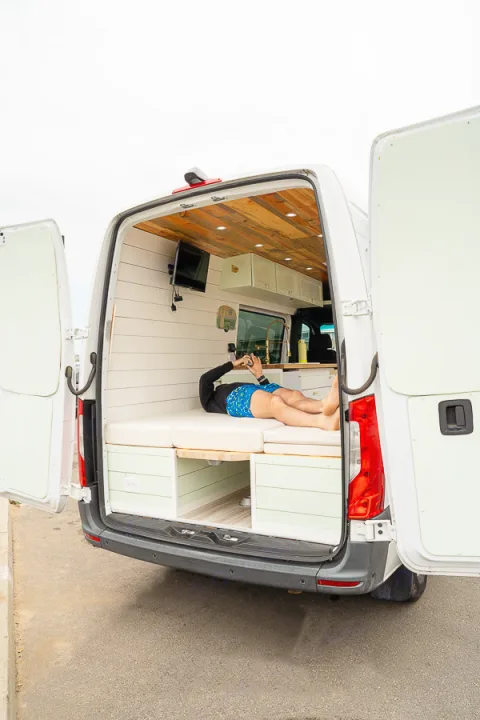 Open back of an RV rental: a person relaxes inside a custom camper van with a cozy bed, TV, and wood-paneled ceiling.. Mercedes-Benz Sprinter 2019