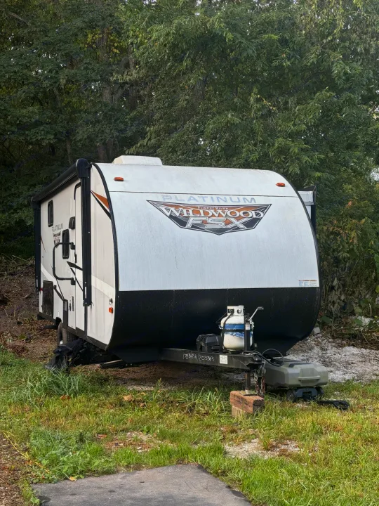 A white & black Platinum Wildwood FSX travel trailer, ideal for an rv rental, is parked outdoors amidst green trees.. Forest River Wildwood FSX Plantium 2022
