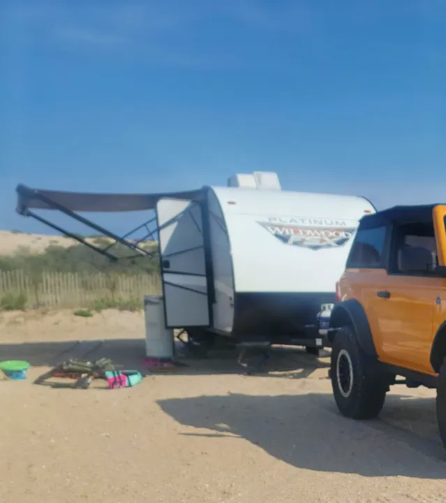 A yellow Bronco tows a white Wildwood FSX RV rental, parked on a sandy beach with its awning extended for beach camping.. Forest River Wildwood FSX Plantium 2022