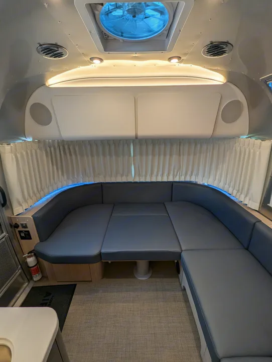 View of a modern RV rental interior featuring a grey U-shaped lounge, convertible bed, ceiling fan, and white curtains.. Airstream Globetrotter 2024