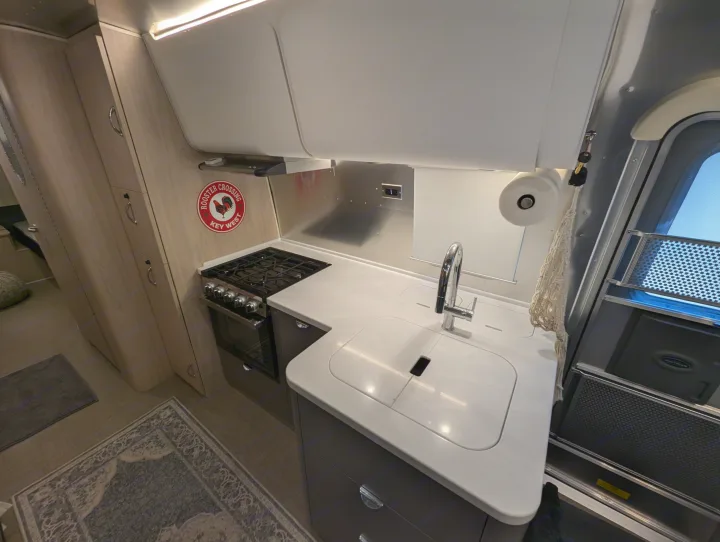 Compact Airstream RV rental kitchen, featuring a stove, sink, and light wood cabinetry on the left.. Airstream Globetrotter 2024