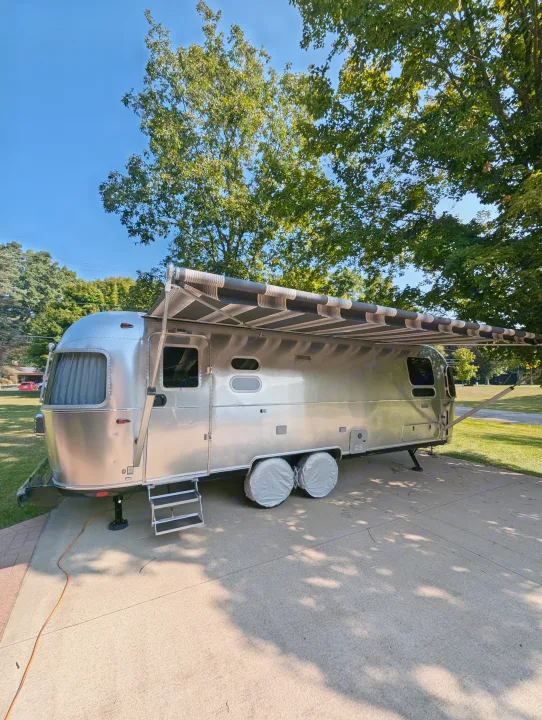 A gleaming Airstream travel trailer, ready for adventure! Awning out, wheels covered, parked on a sunny driveway. Your next rv rental.. Airstream Globetrotter 2024