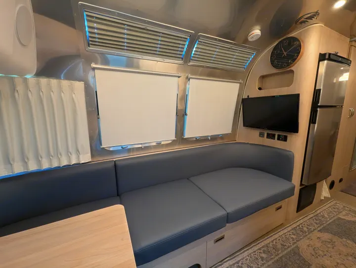 Inside an Airstream-style RV rental: curved metallic walls, a grey dinette, TV, fridge, and light wood cabinetry.. Airstream Globetrotter 2024