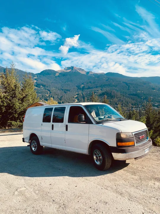 White GMC RV rental van parked amidst stunning mountains and forests under a blue sky. Ideal for your next outdoor adventure.. Gmc Savanna 2016