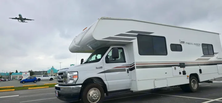 A white Freeland Coachmen RV rental parked, with a low-flying plane overhead, at an airport lot.. Freelander Coachman 2023