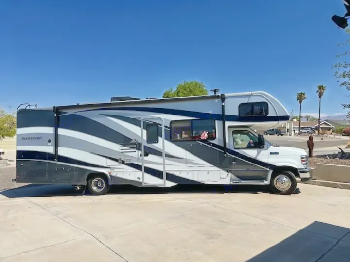 This white and gray Sunseeker Class C RV, perfect for your next rv rental, is parked with its awning extended on a sunny drive.. Forest River Sunseeker 2018