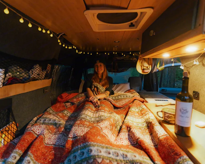 A woman smiles cozily in an RV rental, covered by a patterned blanket with warm string lights above.. Ford Transit 2011