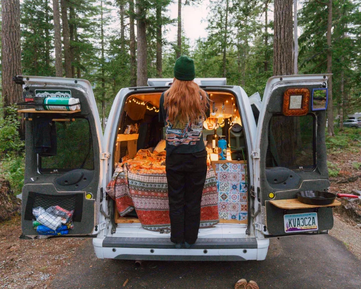 Cozy rv rental setup: Person at open rear of white camper van, showcasing organized interior with bed, kitchen, and lights.. Ford Transit 2011