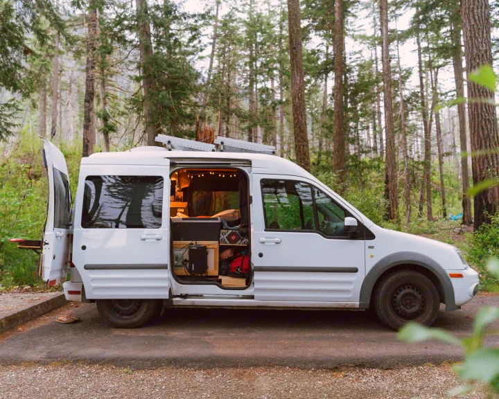 A white Ford Transit Connect camper van, customized for van life with solar panels & interior lights, parked in a forest. Ideal RV rental.. Ford Transit 2011