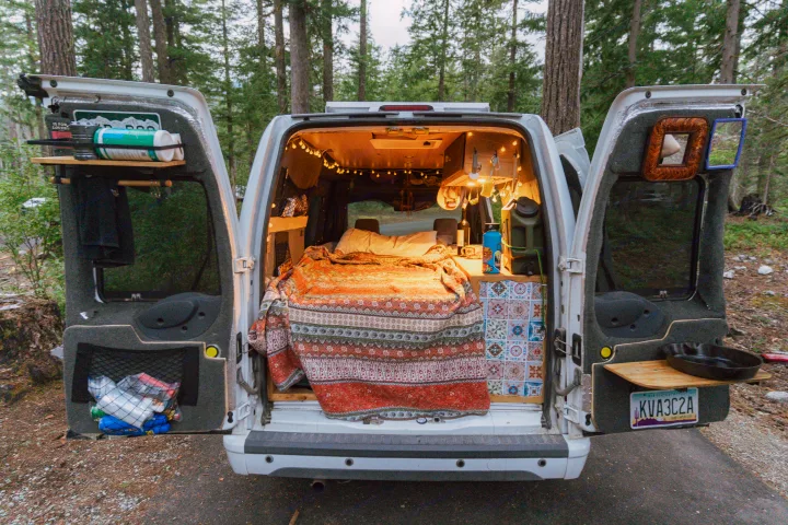 View from rear of a cozy camper van, doors open. Inside: bed, fairy lights, kitchen setup. Ideal for an RV rental outdoor adventure.. Ford Transit 2011