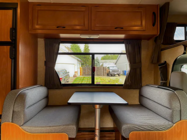 Comfy RV rental dinette featuring wood cabinets, gray benches, a table, and a view outside through the large window.. Thor Majestic 2015