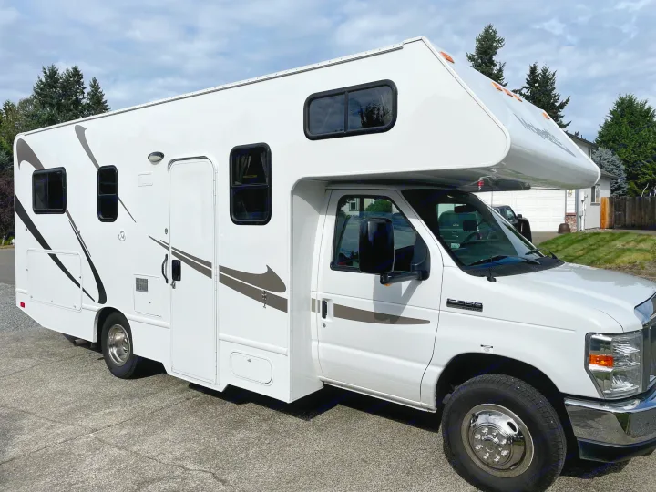 A white Class C RV rental, featuring brown side decals, parked on a paved surface on a cloudy day.. Thor Majestic 2015