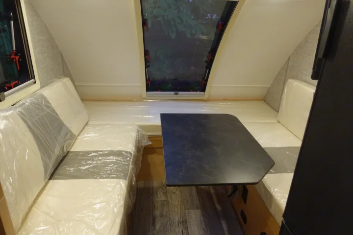 Bright RV rental interior showing dinette benches with plastic-wrapped cushions, a central table, and dual windows.. Little Guy Mini-Max 2024