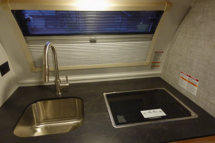 RV rental kitchen: dark countertop with a sink, pull-down faucet, Dometic cooktop, and window blinds.. Little Guy Mini-Max 2024