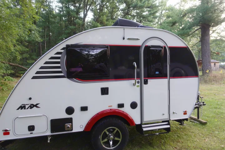 A white and black Mini Max teardrop camper, ideal for an RV rental, is set against a lush green forest.. Little Guy Mini-Max 2024