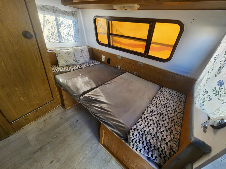 Cozy RV rental interior featuring a bed with patterned cushions, wooden walls, and windows with amber shades.. Fleetwood Wilderness 1991