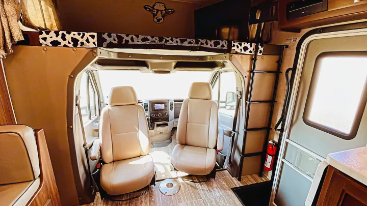 Cozy RV rental interior with front cockpit, over-cab bed with cow print, ladder, and fire extinguisher near the door.. Forest River Sunseeker MBS 2019