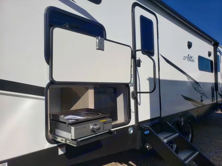 An RV rental's side view: a white travel trailer with an open compartment showcasing an integrated Greystone griddle, plus entrance steps.. East to West Alta 2024