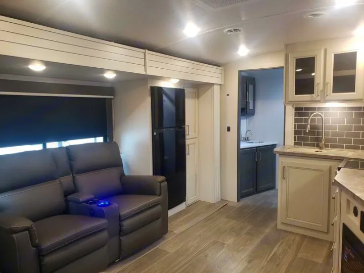 Modern RV rental interior: living area with dark recliners, a black fridge, light grey kitchen cabinets, and subway tile backsplash.. Keystone RV Cougar 2024