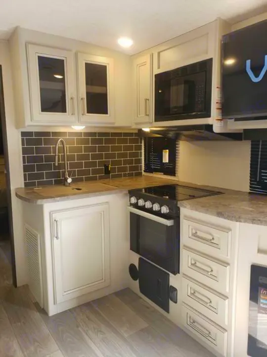 Modern RV rental kitchen with light cabinets, dark subway tile backsplash, sink, stove, microwave, and wood-look floor.. Keystone RV Cougar 2024