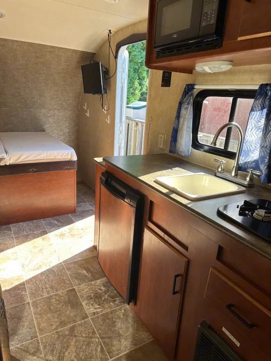 Bright rv rental interior showing a compact kitchen with sink, stovetop, fridge, microwave, and a cozy bed area.. Forest River Salem Cruise Lite 2017