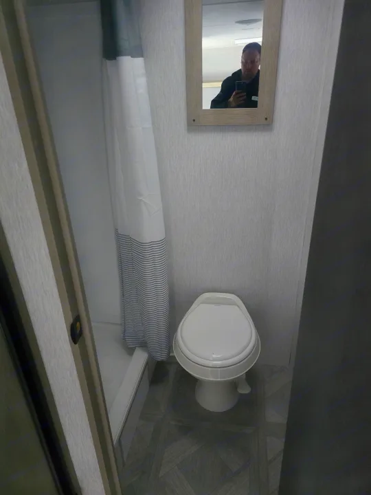 Inside an RV rental bathroom: a white toilet, striped shower curtain, and a mirror reflecting the person taking the photo.. Jayco Jay Flight SLX 2024