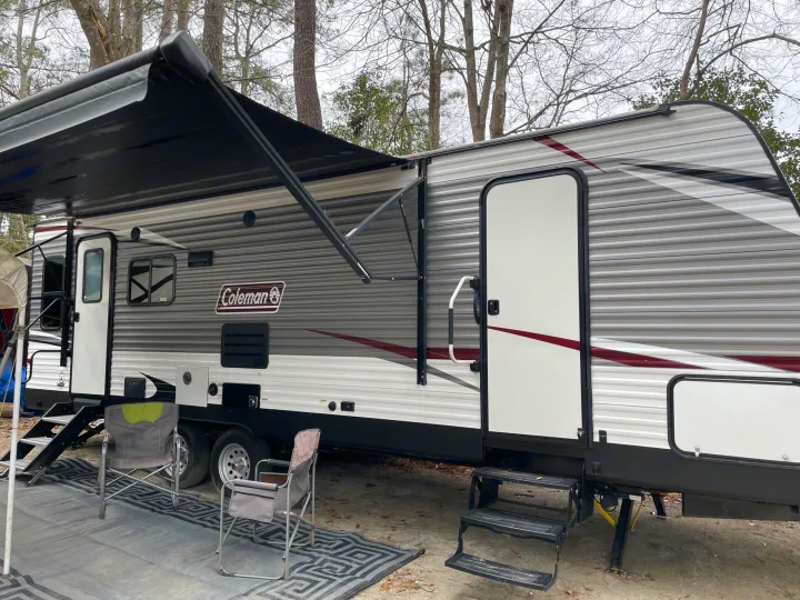 Side view of an RV rental parked at a campsite, with an extended awning, steps, and outdoor chairs set up.. Coleman Lantern 2019