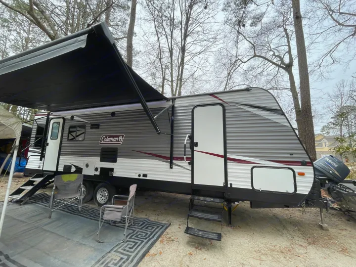 A grey Coleman RV rental is perfectly set up at a wooded campsite, featuring an extended awning and comfortable outdoor chairs on a rug.. Coleman Lantern 2019