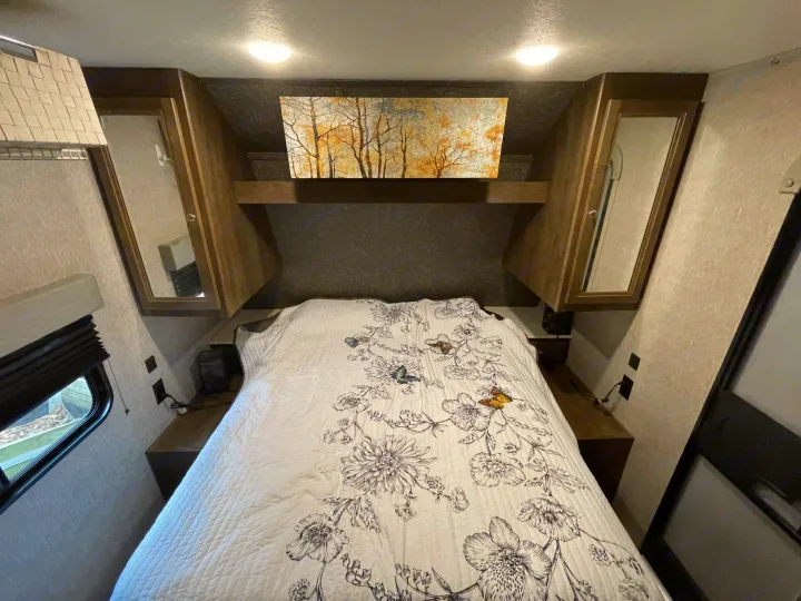 Spacious RV rental bedroom with a white floral quilt, built-in mirrored cabinets, and tree artwork. Outlets visible for charging.. Coleman Lantern 2019