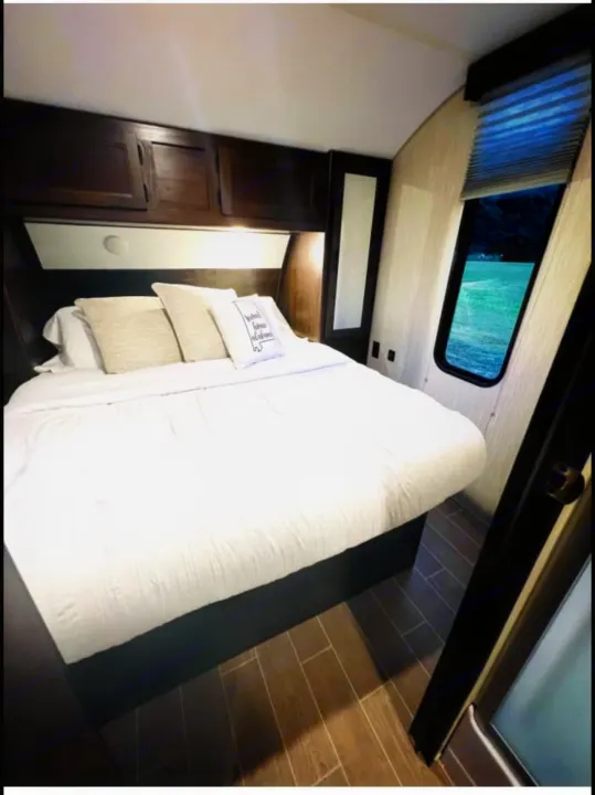 Cozy RV rental bedroom featuring a large bed with white linens, dark wood cabinets, and a window with a scenic lake view.. Forest River Cherokee Grey Wolf 2020