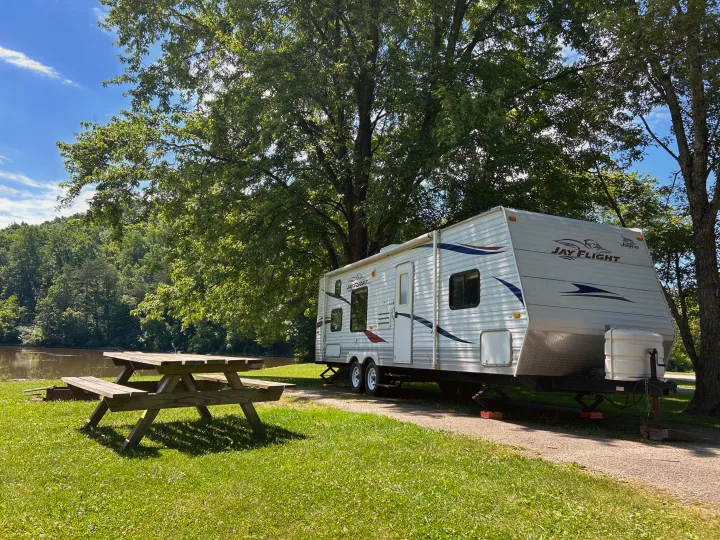 A white Jay Flight RV rental is parked at a scenic lakeside campsite with a picnic table and trees.. Jayco Jay Flight 2010