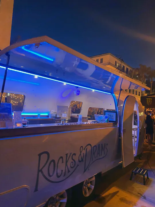 Stylish RV rental transformed into a mobile bar, "Rocks & Dramas," glowing with blue lights at an evening event.. Airstream custom 1973