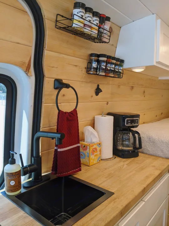 Inside this cozy RV rental, a compact kitchen offers wood paneling, a black sink, coffee maker, and neatly organized spices.. Sprinter Mercedes 2018