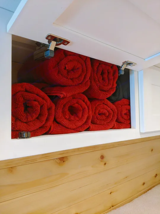 RV rental storage: Neatly rolled red towels in an open overhead cabinet with wood paneling below.. Sprinter Mercedes 2018