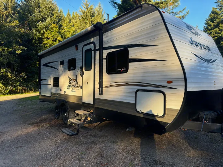 This white & black Jayco travel trailer rv rental is parked on gravel near tall trees, ready for adventure with steps extended.. Jayco Jay Flight SLX Baja Edition 2017