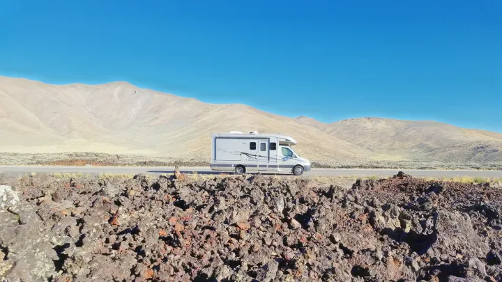 Grey RV rental parked by large lava rock formations, with arid mountains under a clear blue sky in the background.. Coachmen Prism 2009